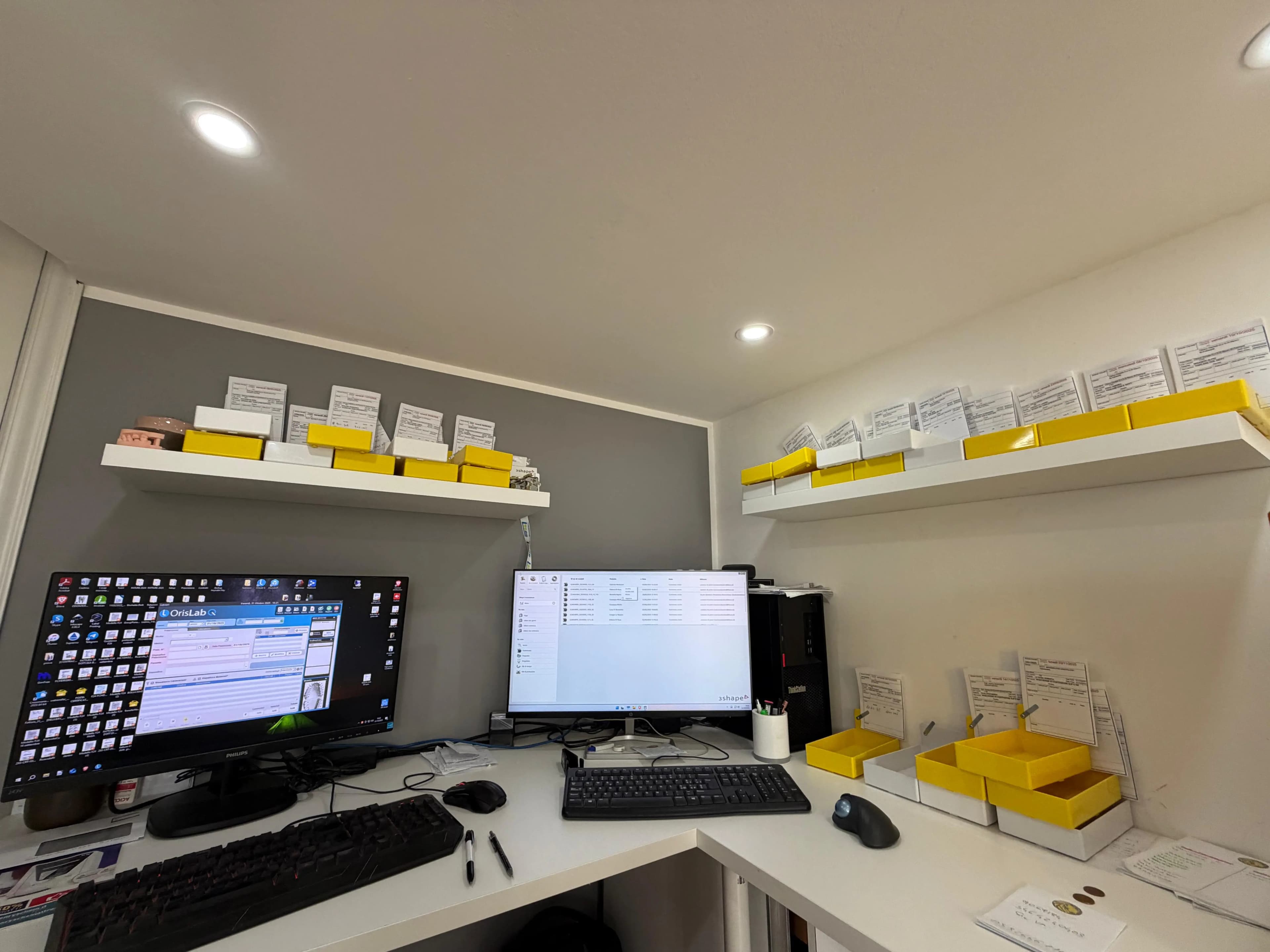 La postazione CAD con il gestionale e 3shape sui due monitor, e le scatole dei casi in lavorazione sulle mensole